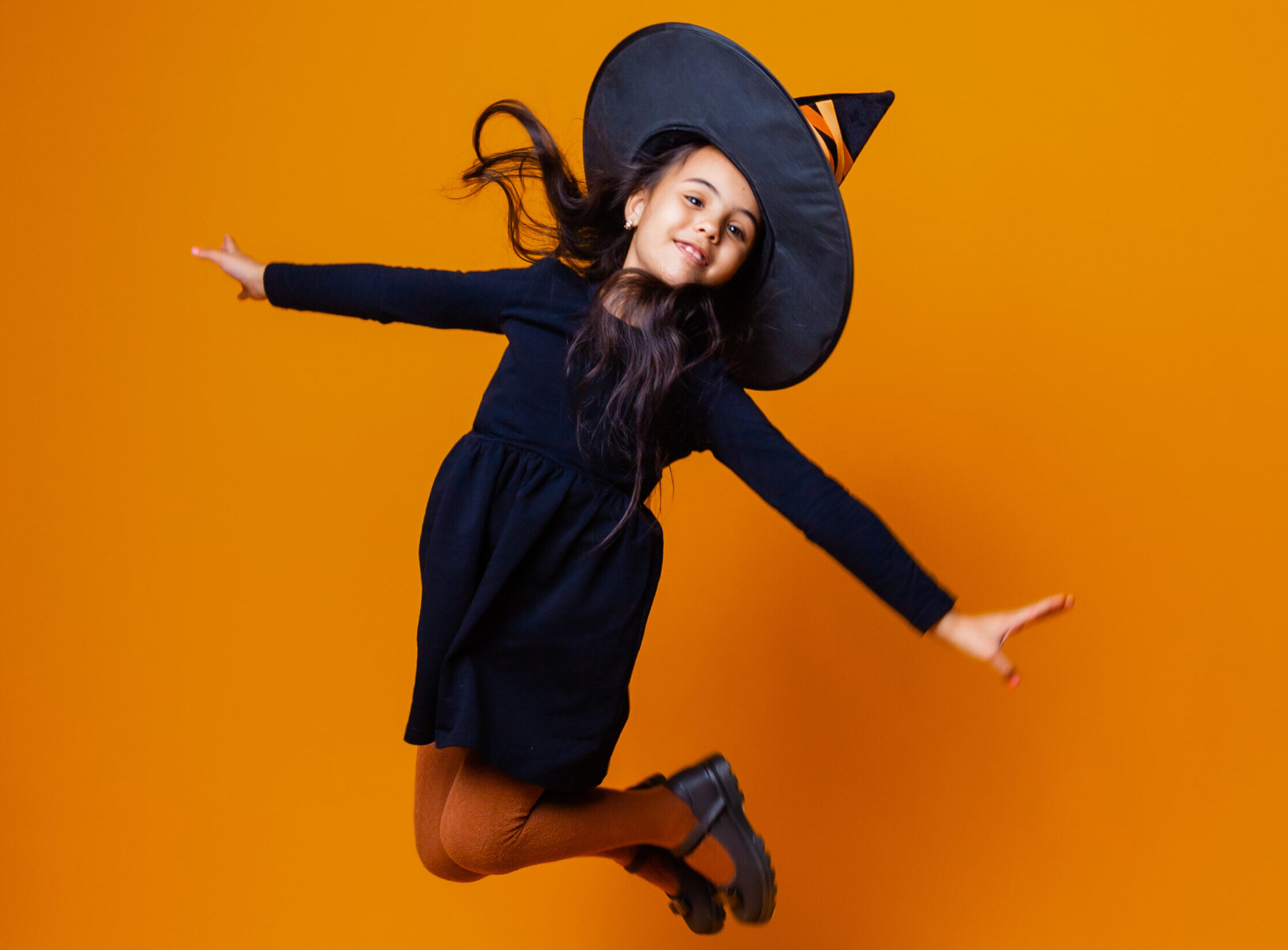 Happy Caucasian girl in a witch costume jumping on an orange background. halloween.