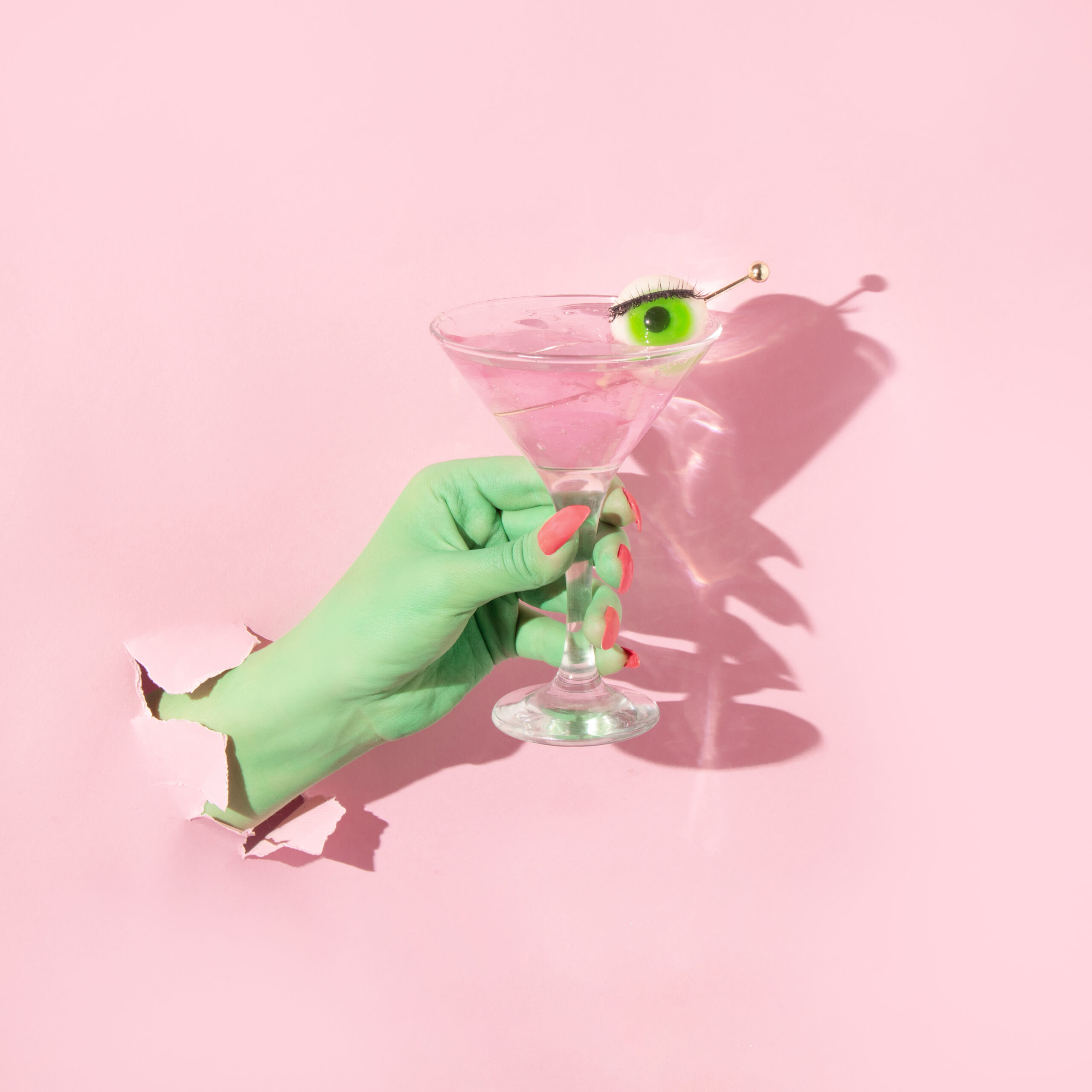 Halloween creative layout with green witch hand with bright pink nails holding martini cocktail glass with eyeball against pastel pink background. Aesthetic season idea. Minimal Halloween concept.